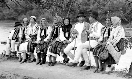 Rumunski seljaci sede pored reke u svom selu na klupi. Tu su i muski i zenski i mladi i stari.