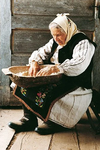 Stara vlajna tj rumunka obučena u narodnu nošnju. Drži korito u krilu i mesi brašno tj priprema hleb